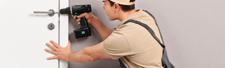 Handwerker in Latzhose und Kappe montiert mit einem Akkuschrauber ein Türschloss an einer weißen Innentür. Er kniet auf dem Boden und hält die Tür mit der linken Hand fest